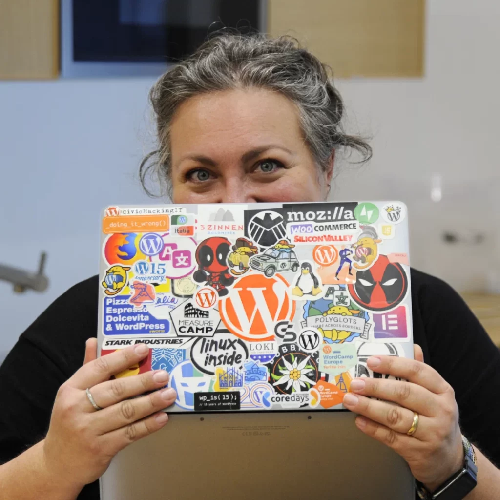 Margherita Pelonara sorride dietro al suo laptop coperto di sticker colorati legati al mondo open source, WordPress e cultura pop.